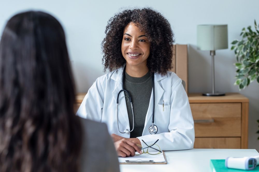 Doctor in white coat smiling, speaking with a patient at a desk.