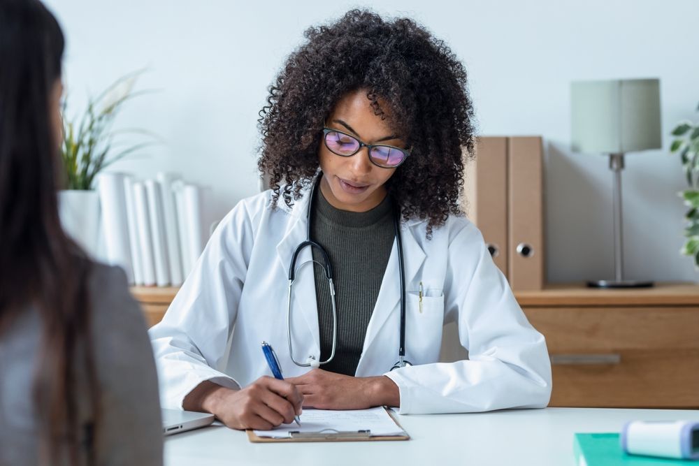 A doctor in a white coat writes on a clipboard while seated at a desk, facing a patient.
