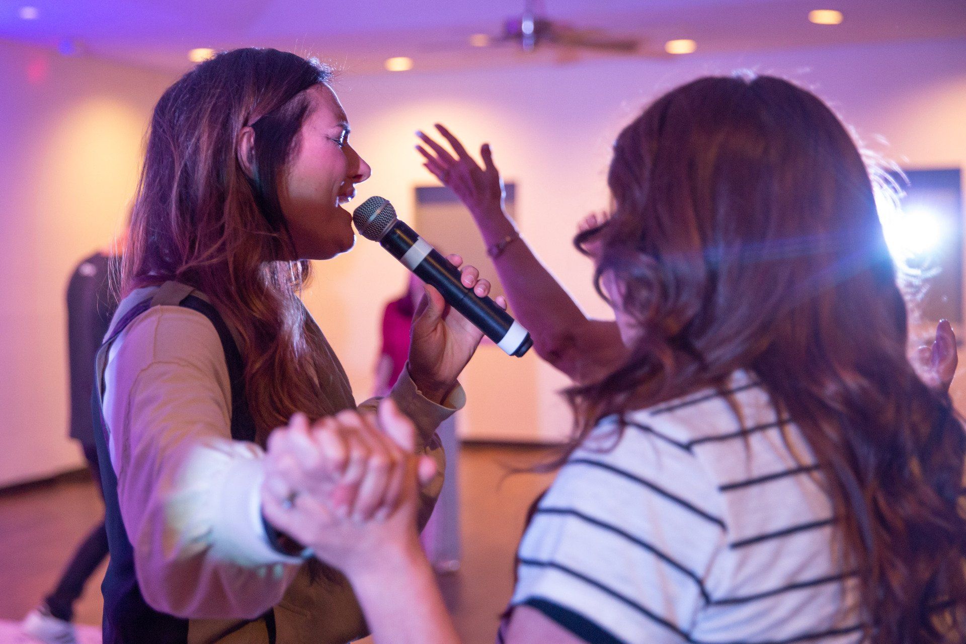 Two women are singing into microphones while holding hands in a room.