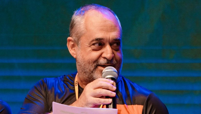 Man speaking into a microphone. He has a gray beard and is wearing a black shirt with orange accents.