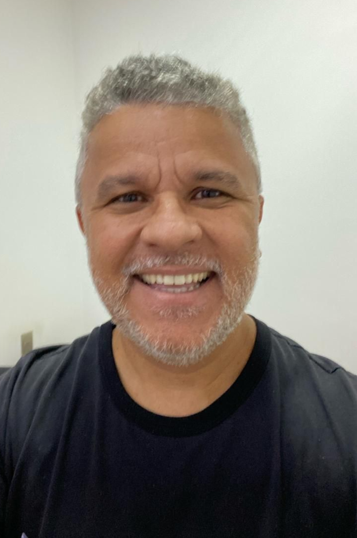 Homem de cabelos e barba grisalhos sorri, vestindo uma camisa preta, em uma sala bem iluminada.