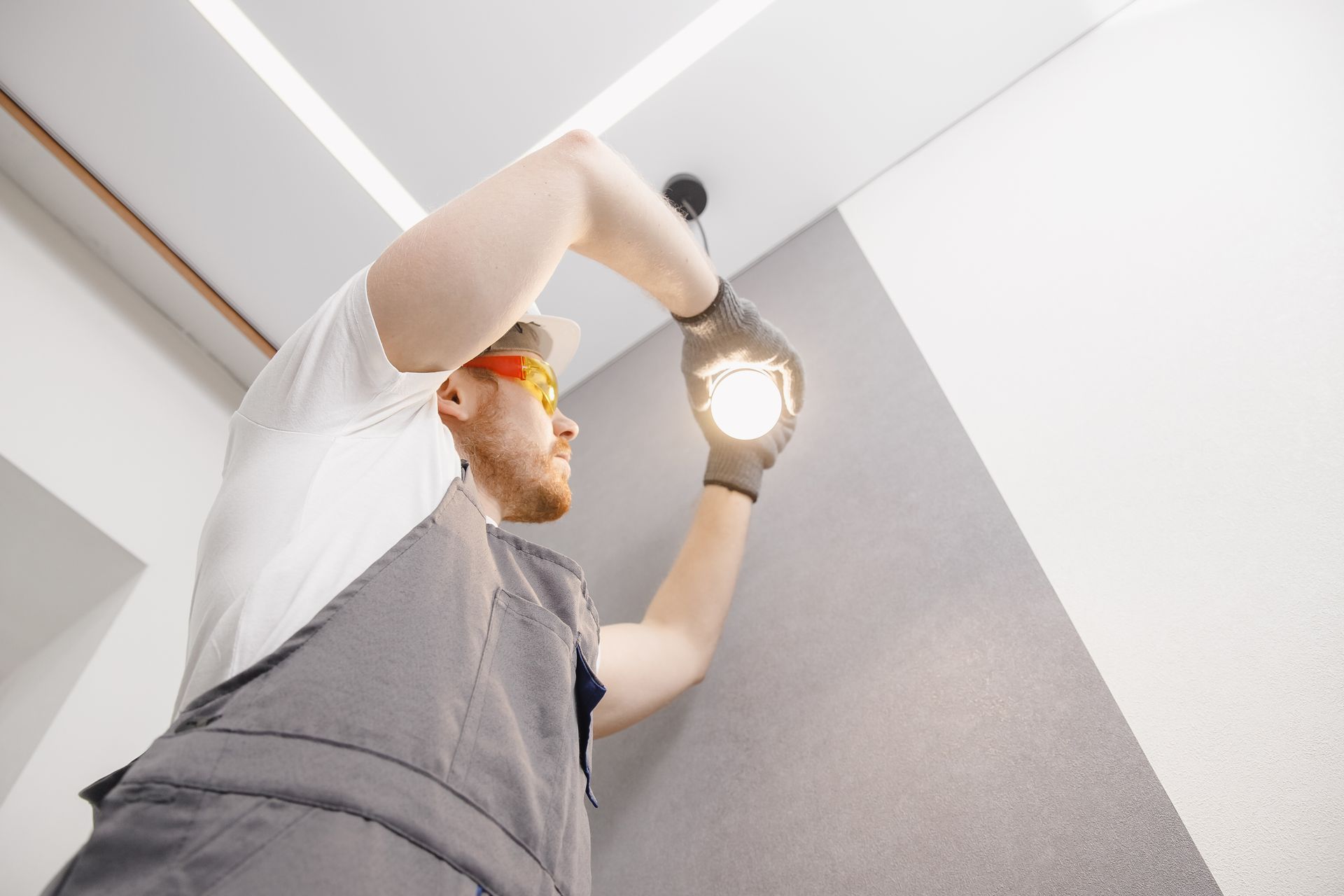A worker in grey overalls and safety glasses installs a bright light bulb into a ceiling fixture.