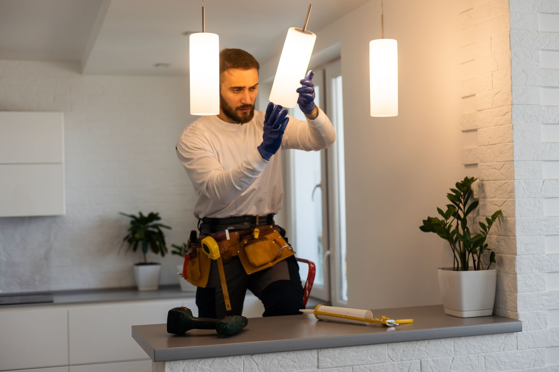 A worker in work pants and a tool belt adjusts a hanging pendant light in a brightly lit, modern kitchen.