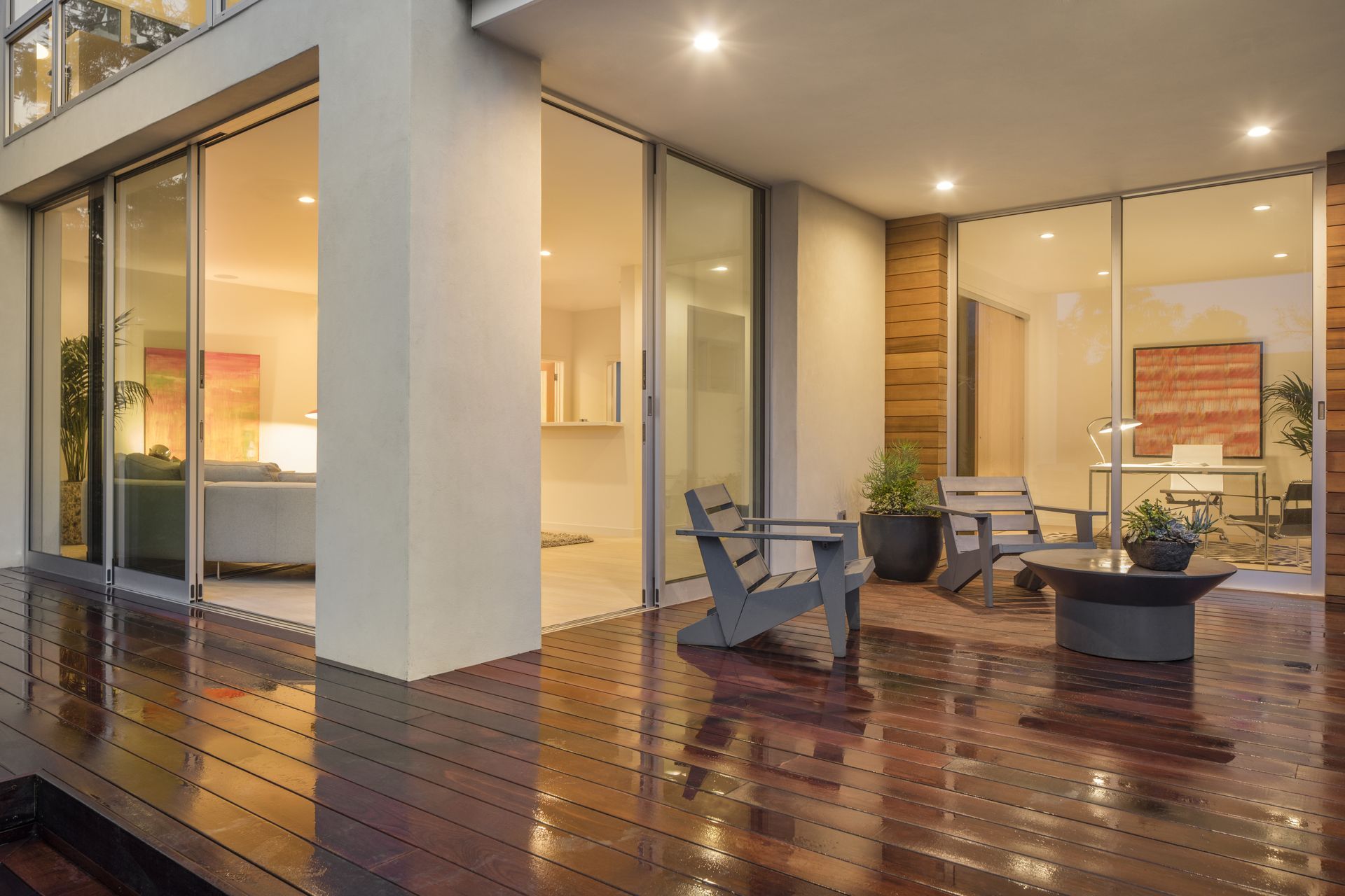 A modern patio with dark wood flooring, two grey chairs, and a small table, seen through large glass sliding doors.