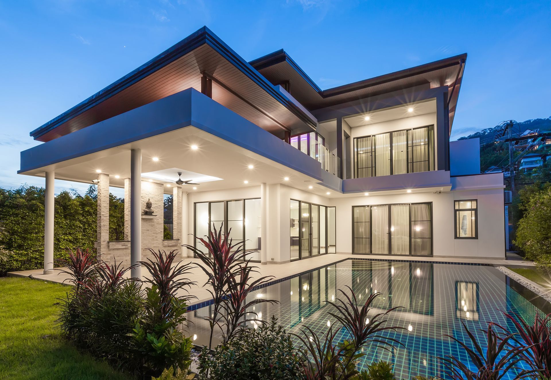 Modern two-story villa at twilight, featuring a white exterior, large sliding glass doors, and an illuminated pool.