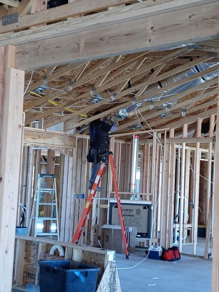 A construction worker stands on a ladder, installing electrical wiring in the ceiling of a wood-framed building.