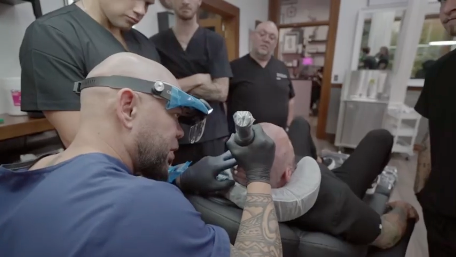 A group of men are watching a man get a tattoo on his arm.