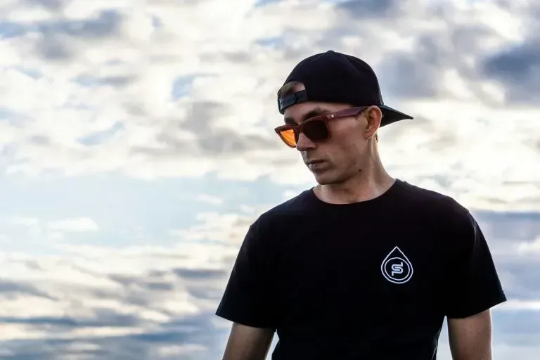 A man wearing sunglasses and a baseball cap is standing in front of a cloudy sky.