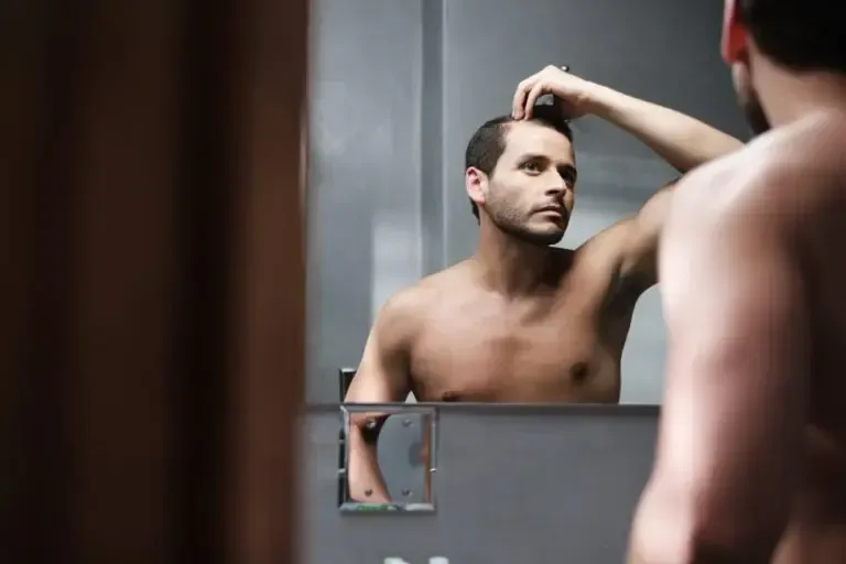 A shirtless man is looking at his reflection in a bathroom mirror.