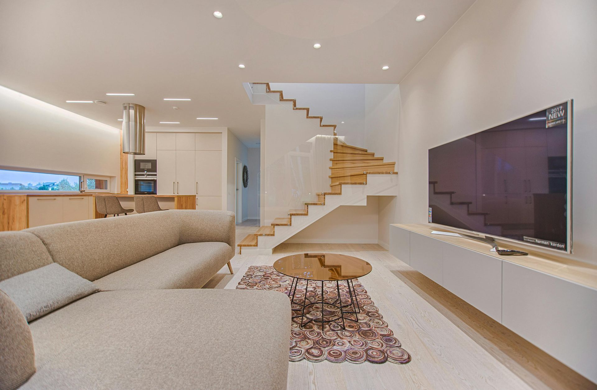 Modern living room with light wood floors, a beige sectional, and a floating TV stand. A staircase leads upwards.