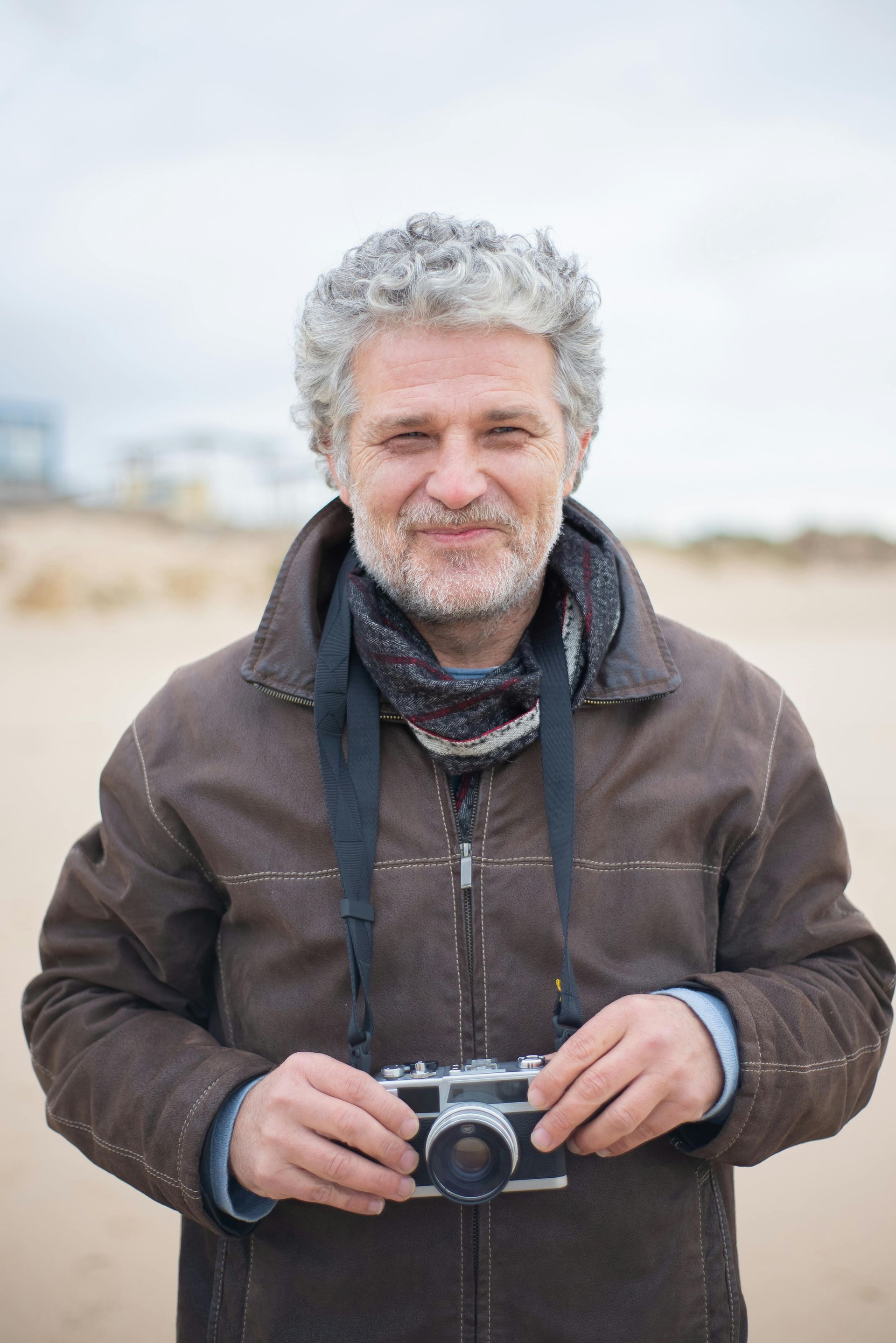 Man with gray curly hair, brown jacket, scarf, and camera outdoors, smiling.