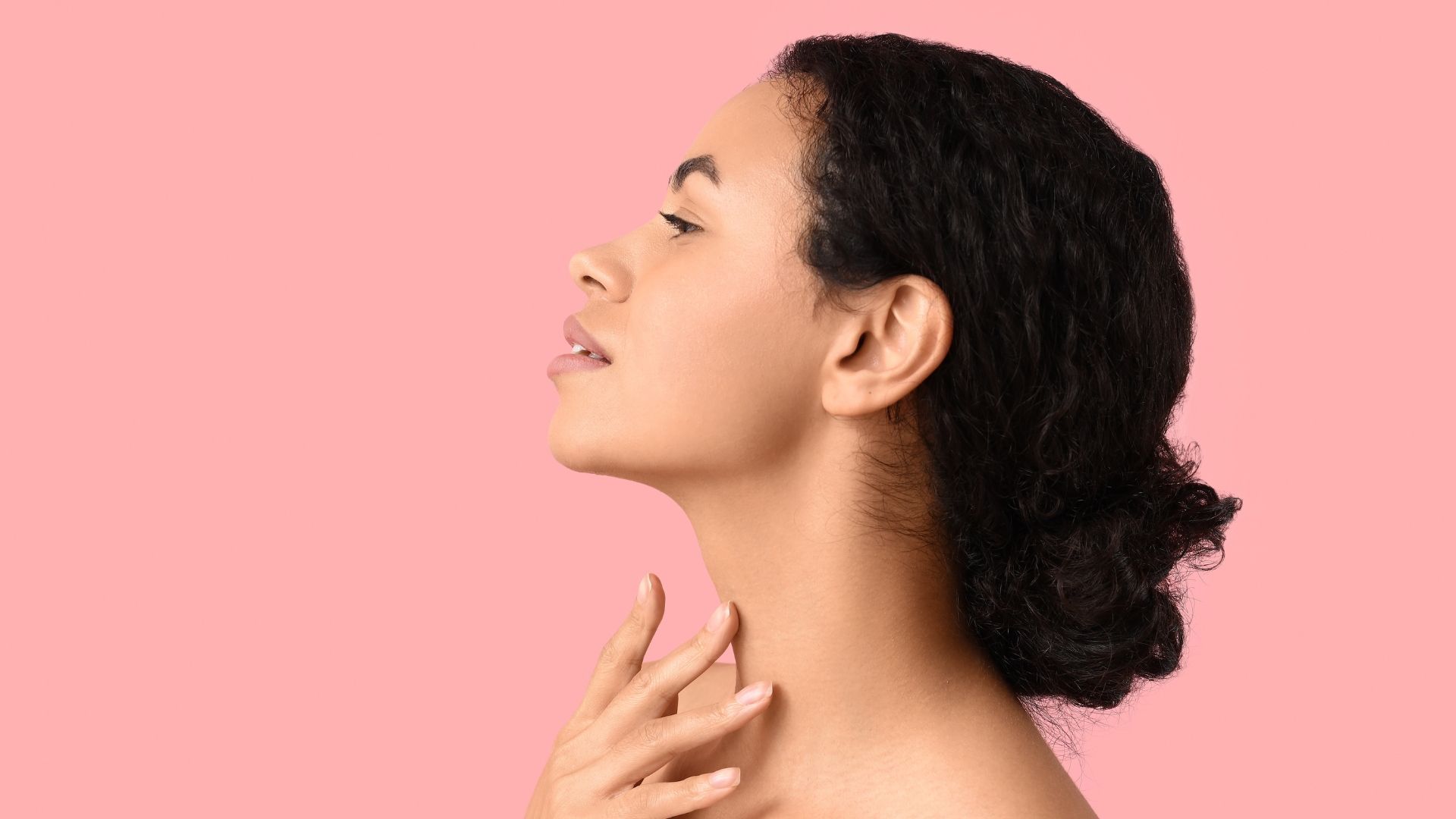 Profile of a woman with dark hair touching her neck against a pink background