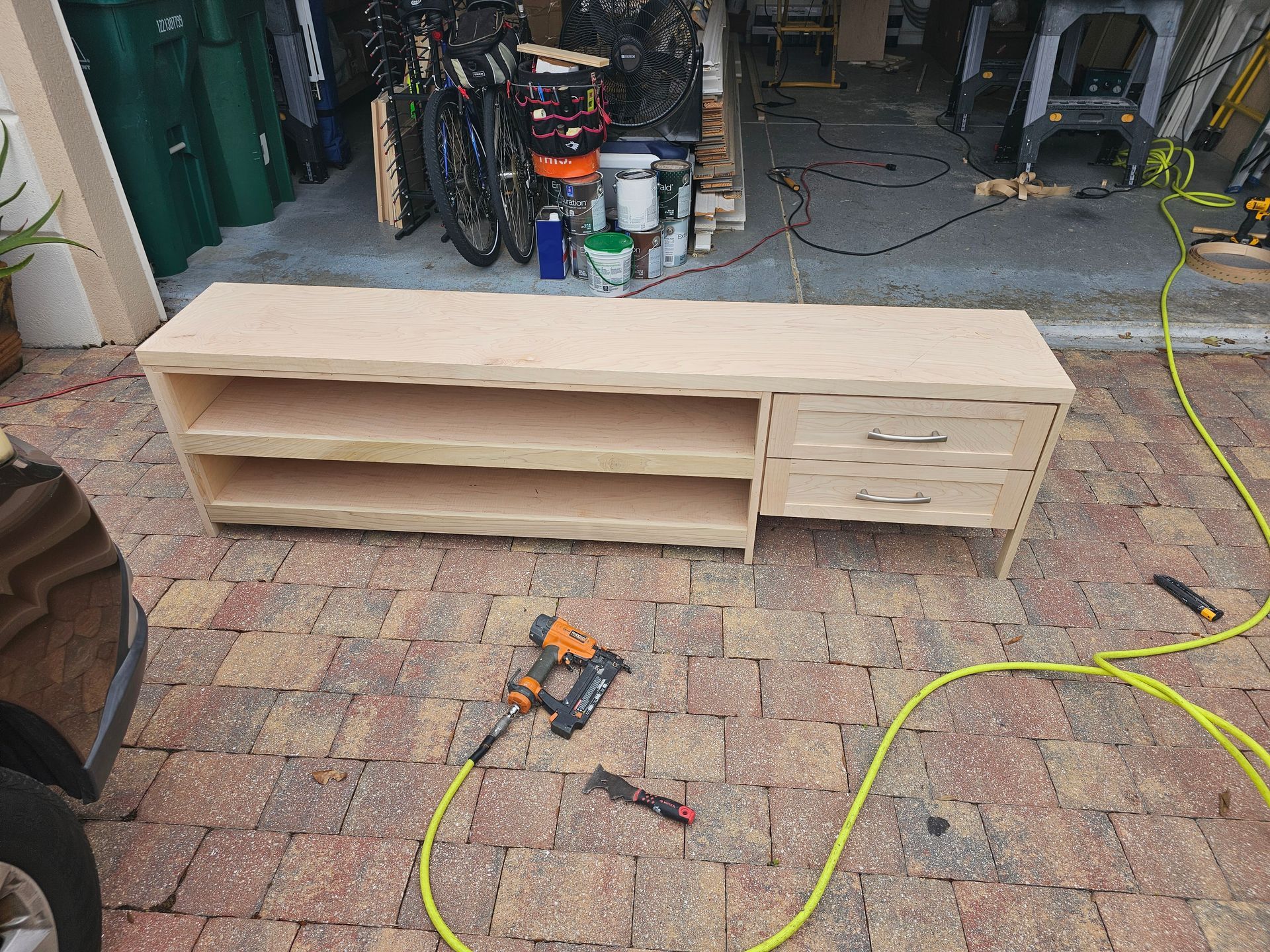 A light wood cabinet with shelves and drawers sits on a brick patio, with tools and garage in the background.