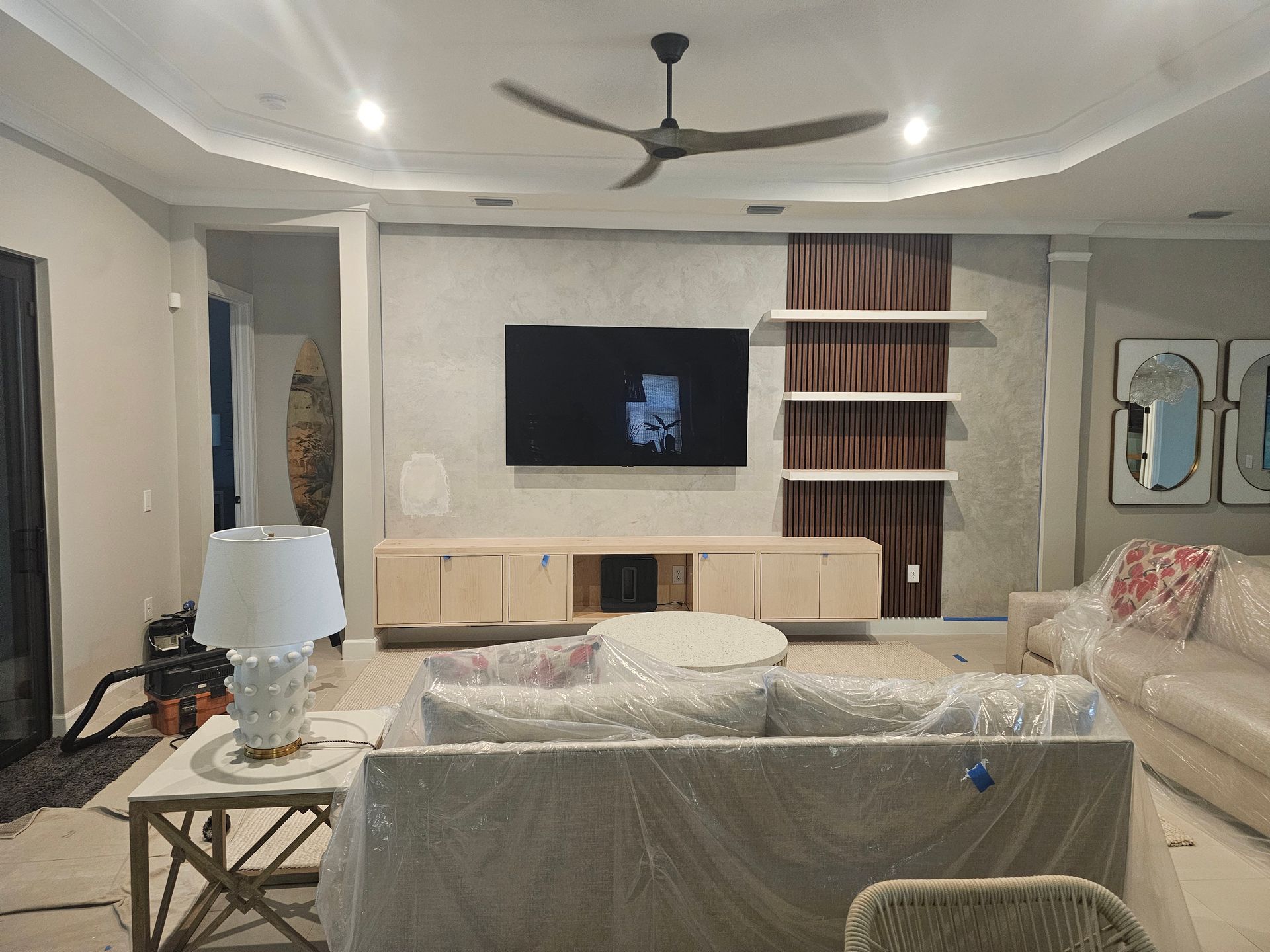 Living room with TV, light wood console, and couch covered in plastic. Ceiling fan, shelves, and lamp visible.