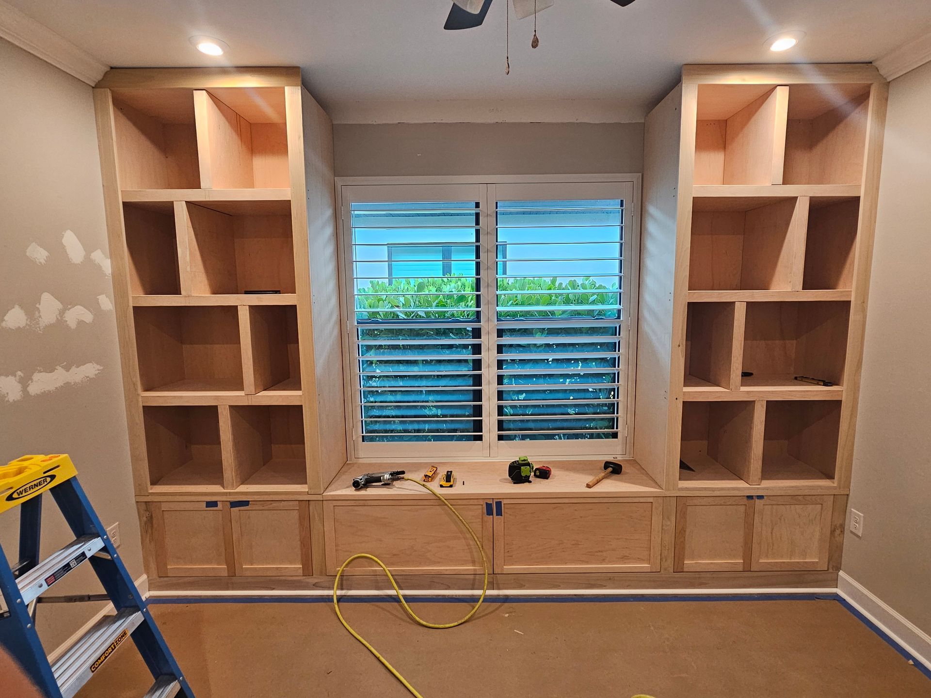 Built-in wooden bookshelves flanking a window with a bench seat, unfinished construction in a room.