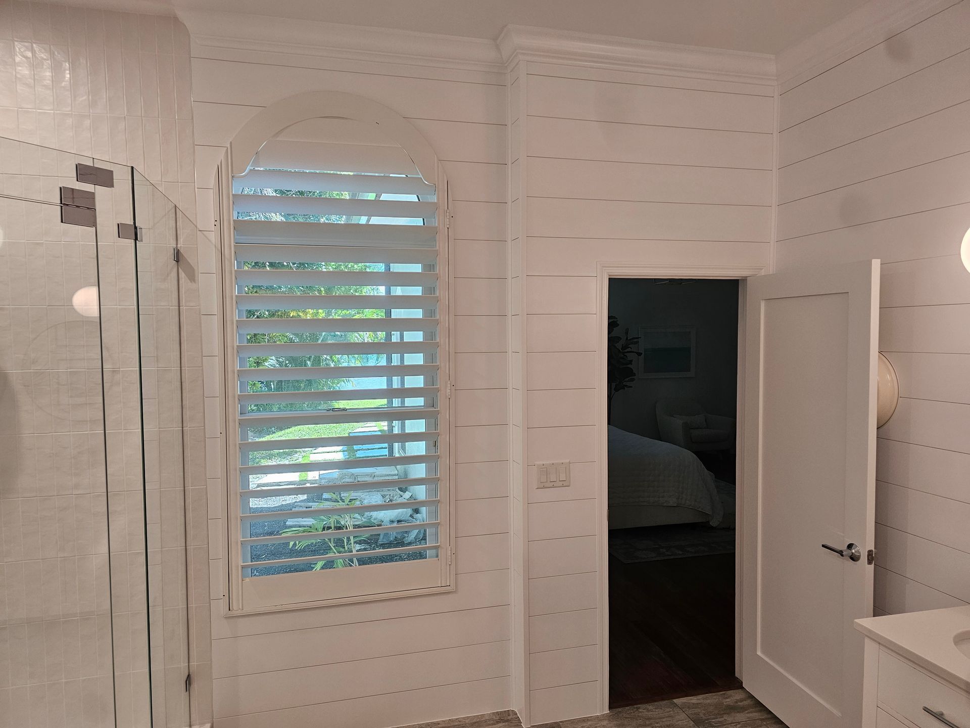 Bathroom interior with arched window, white shutters, open door to bedroom.
