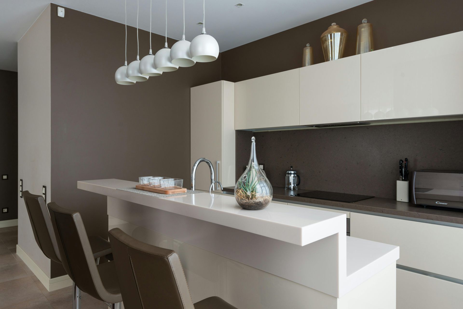 Modern kitchen with a white countertop bar, brown chairs, and white pendant lights.