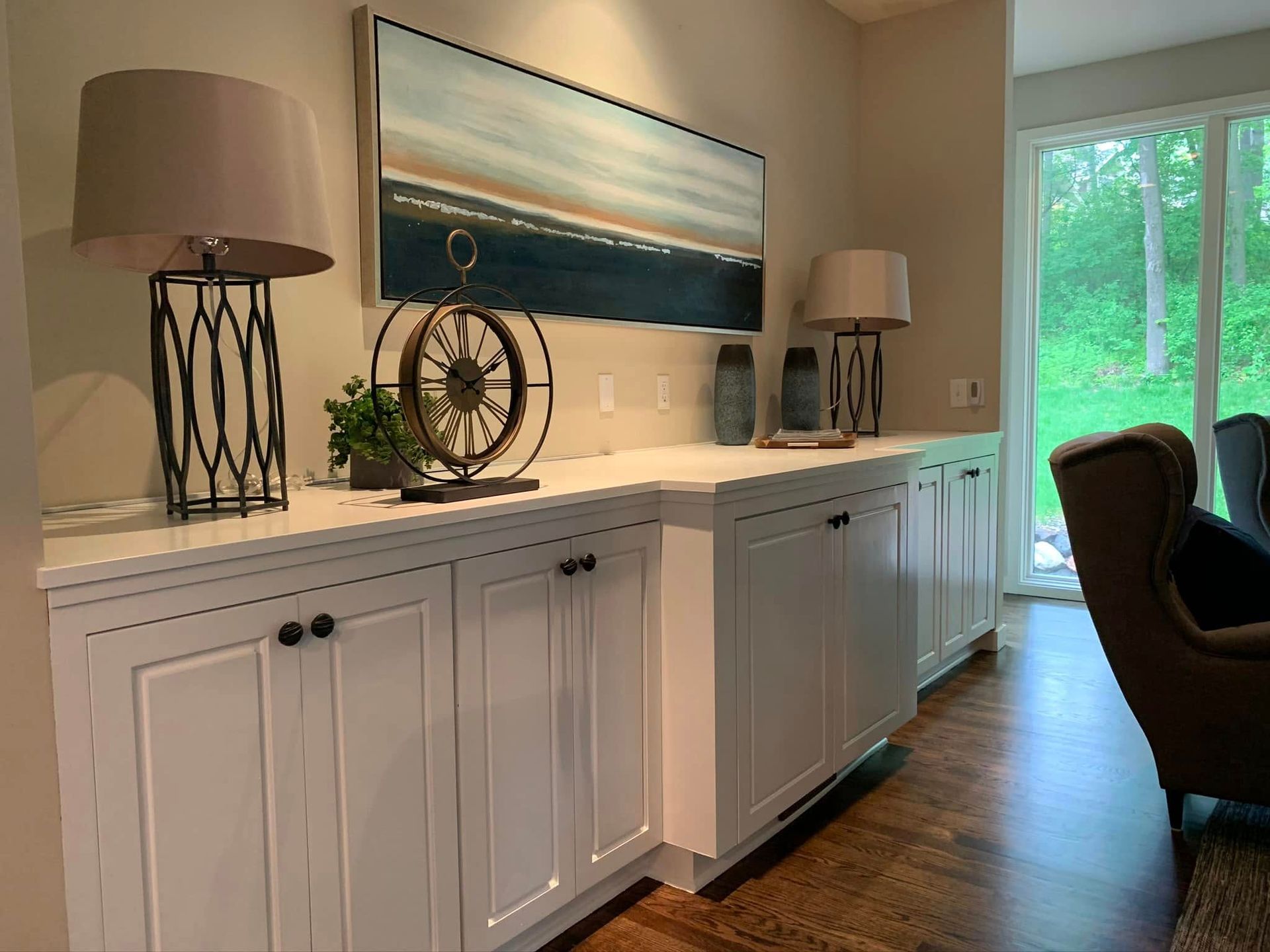 A living room with white cabinets , a chair , lamps , a painting on the wall and a large window.
