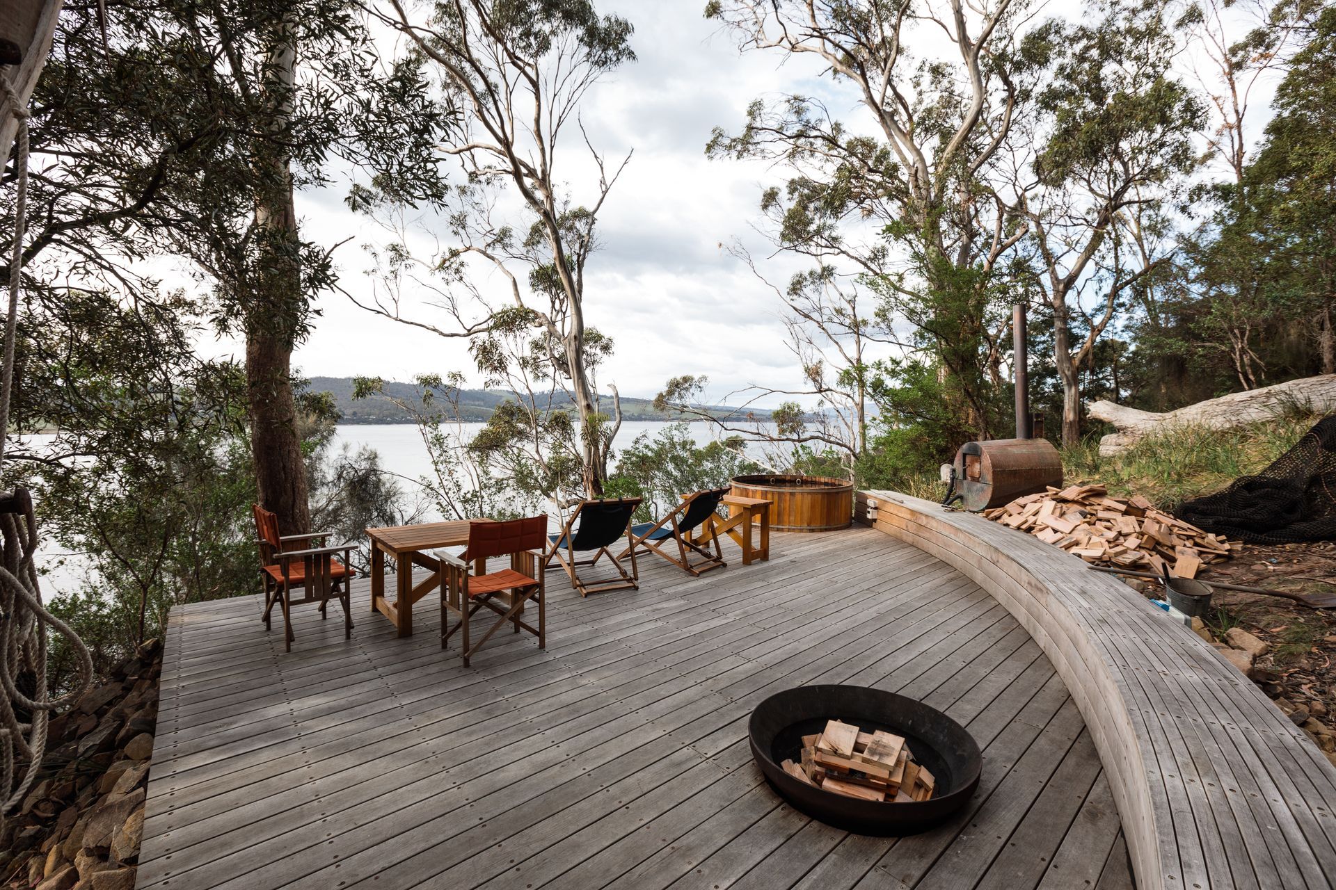 A wooden deck with a fire pit and chairs overlooking a body of water.