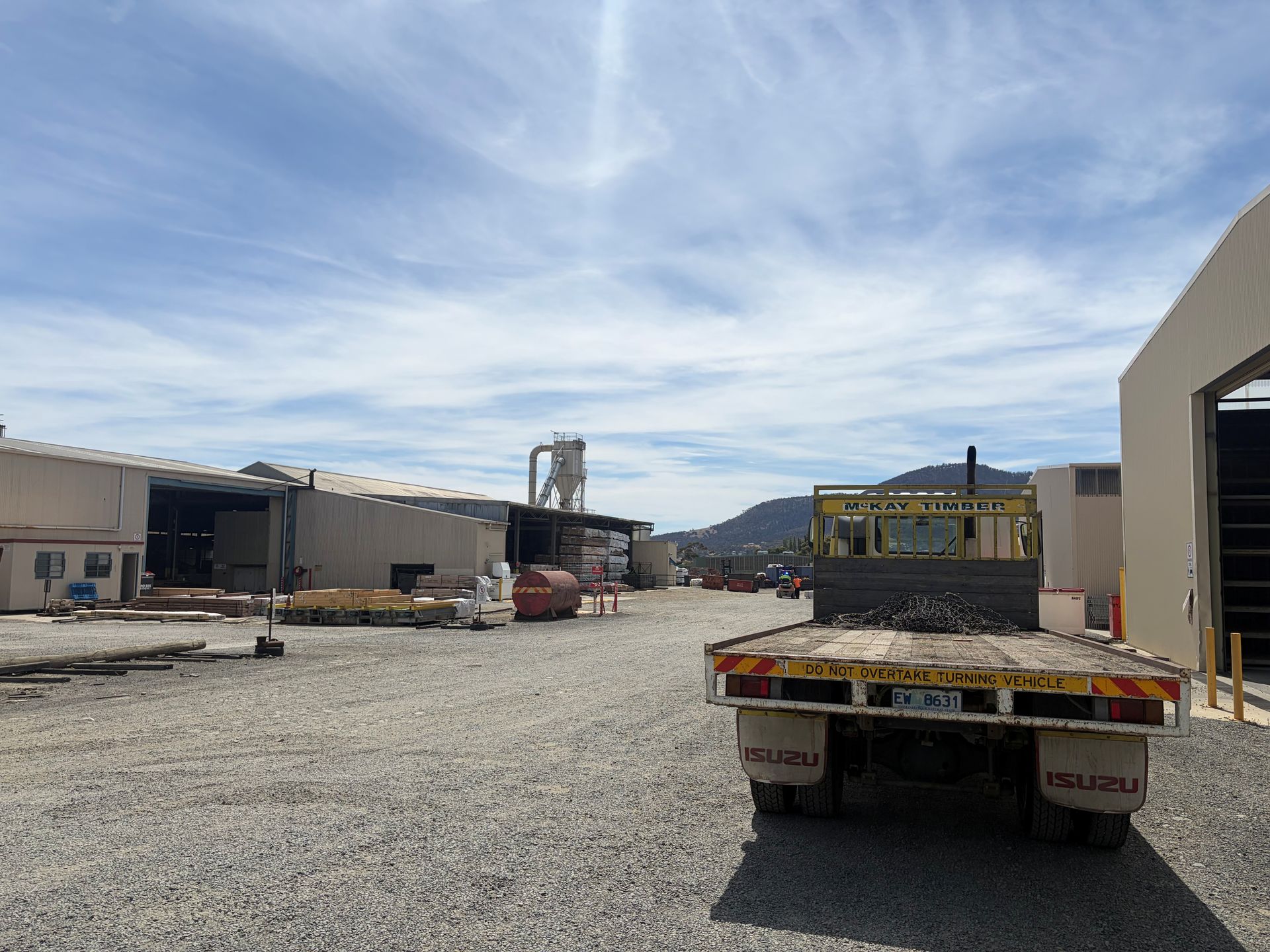 A flatbed truck parked on a gravel lot near industrial buildings under a cloudy sky.