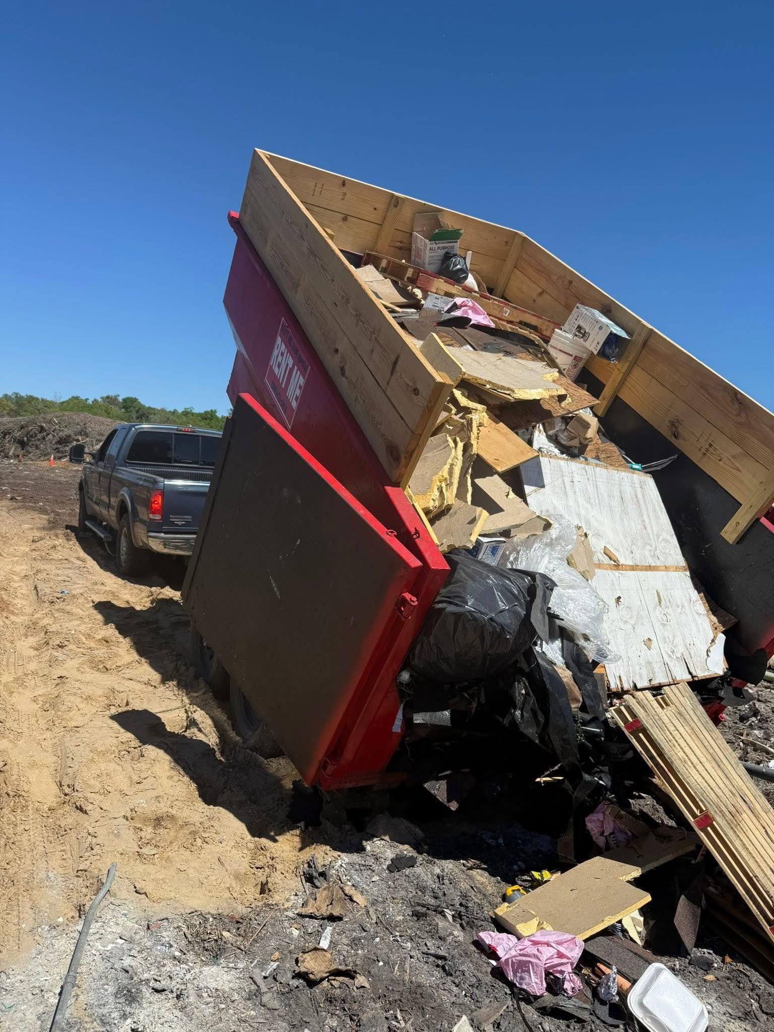 Red dumpster filled with debris, tipped over on dirt, with a truck.