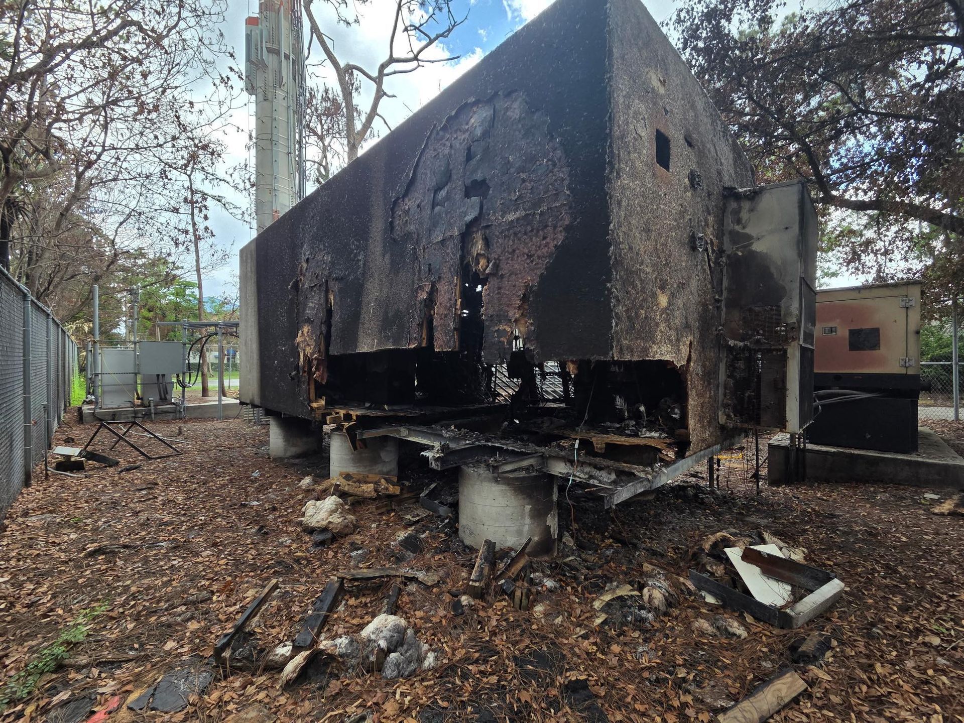 Charred, burnt-out concrete structure with exposed metal. Debris and fallen leaves surround the base, fence in the background.