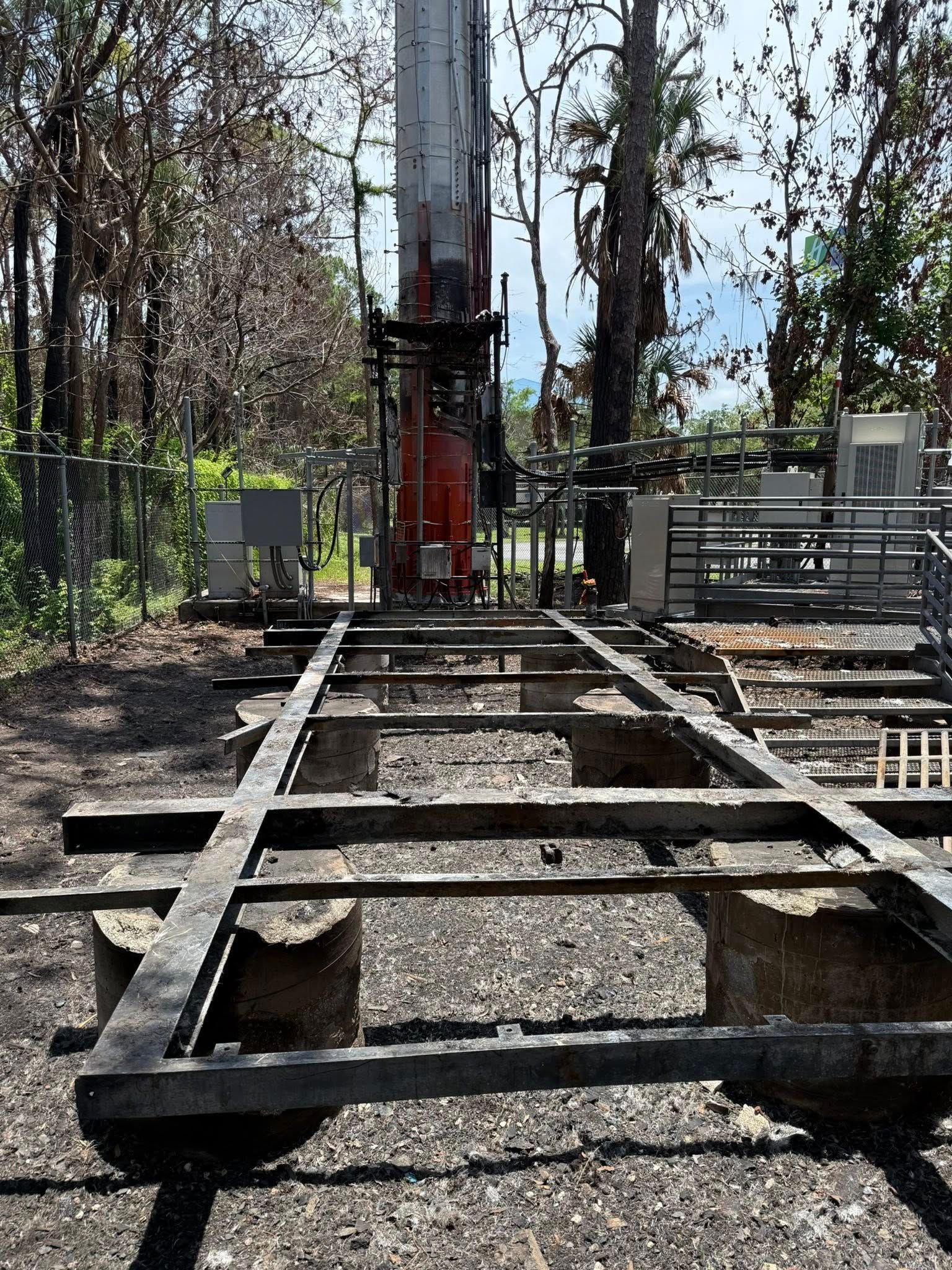 Metal frame on concrete supports beneath a tall communication tower in a wooded area.