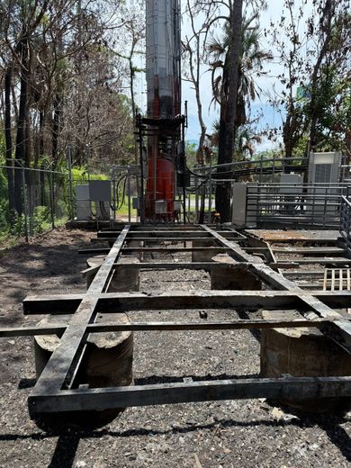 Metal frame on concrete supports beneath a tall communication tower in a wooded area.