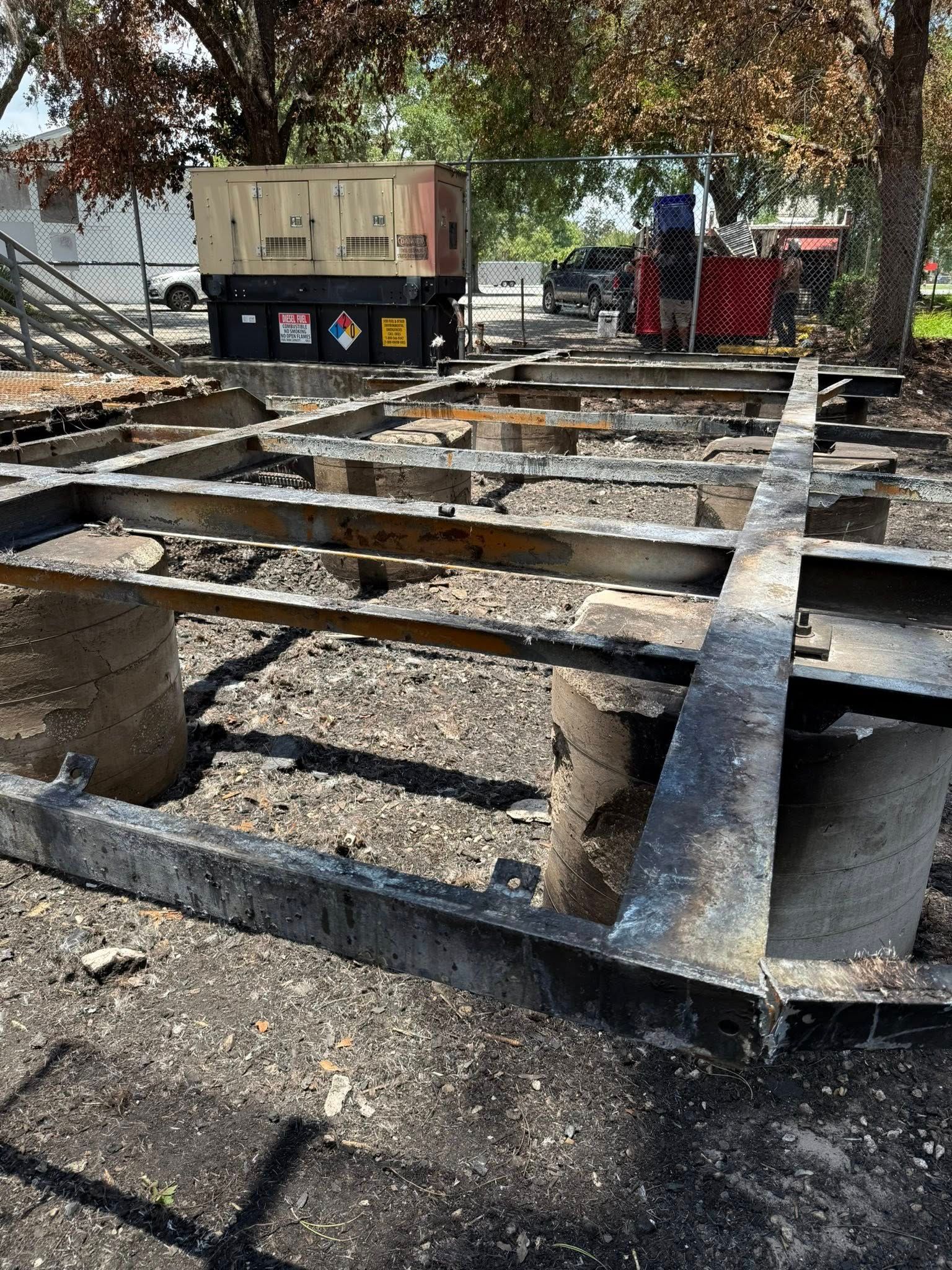 Charred wooden structure on dirt ground, a generator and vehicles in the background.