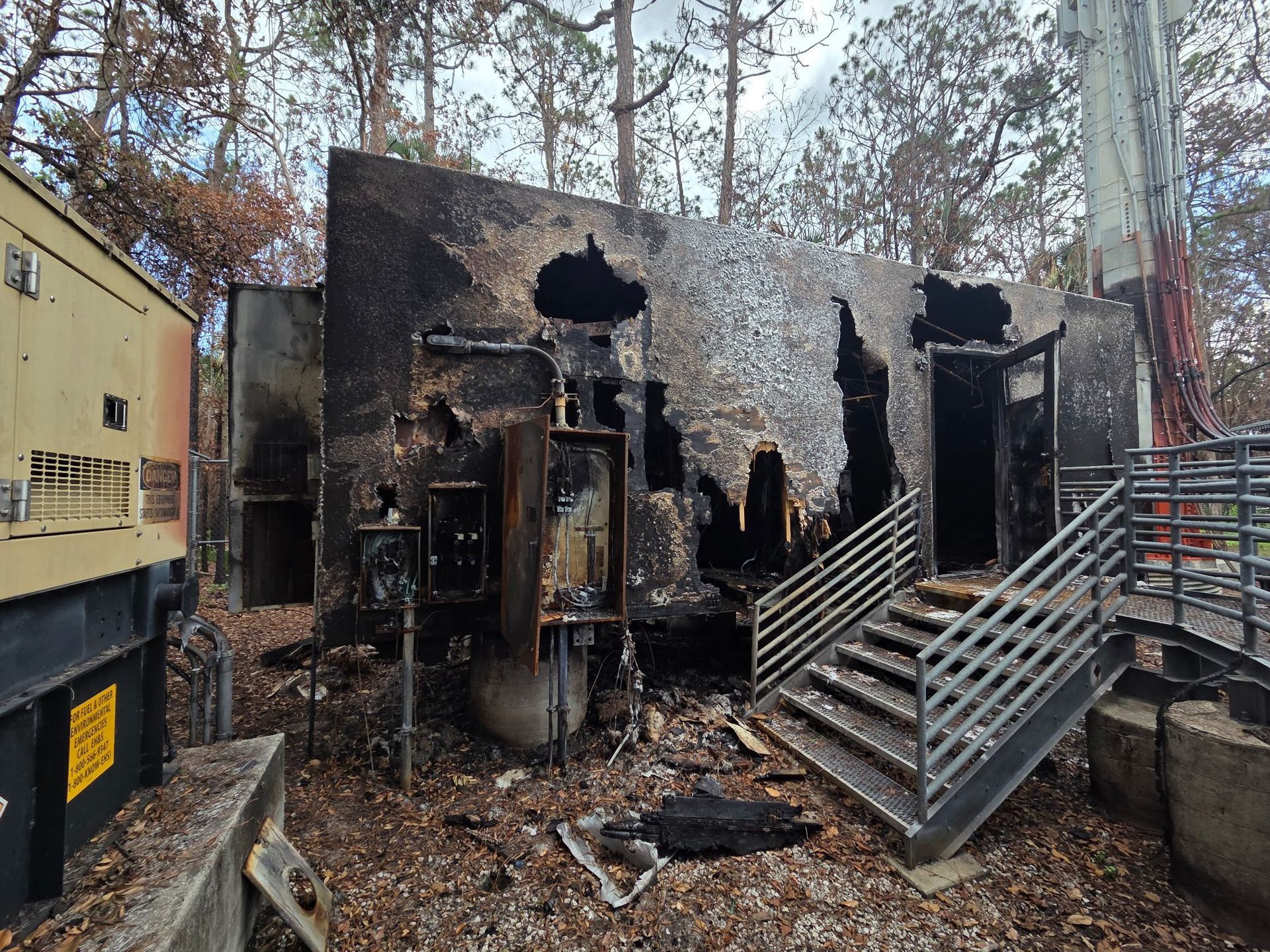 Burned electrical building with stairs, charred exterior, set in a wooded area.