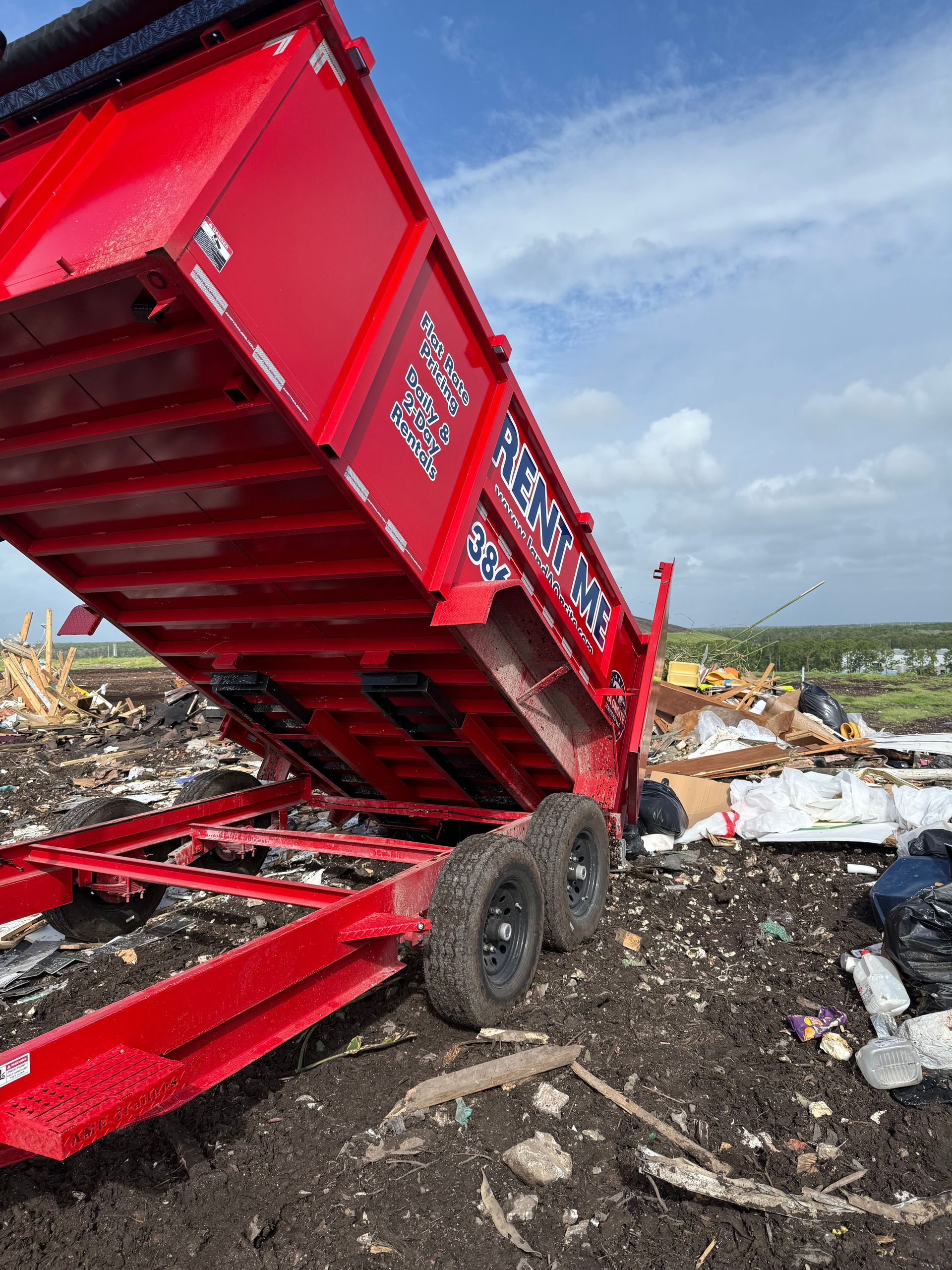 Red dump trailer, raised, unloading debris at a landfill, blue sky.