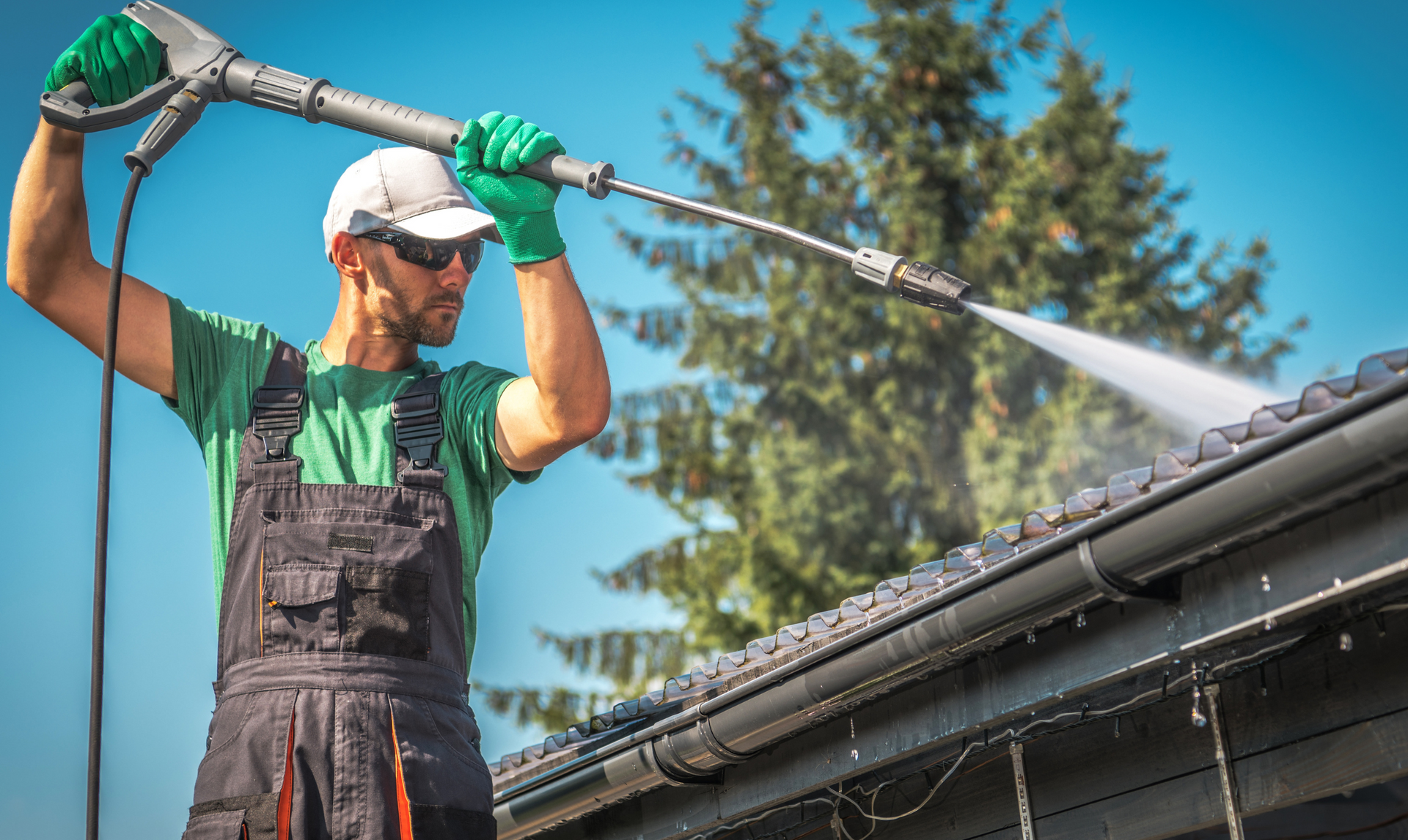 Man in blue helmet pressure washing a red tile roof on a sunny day.