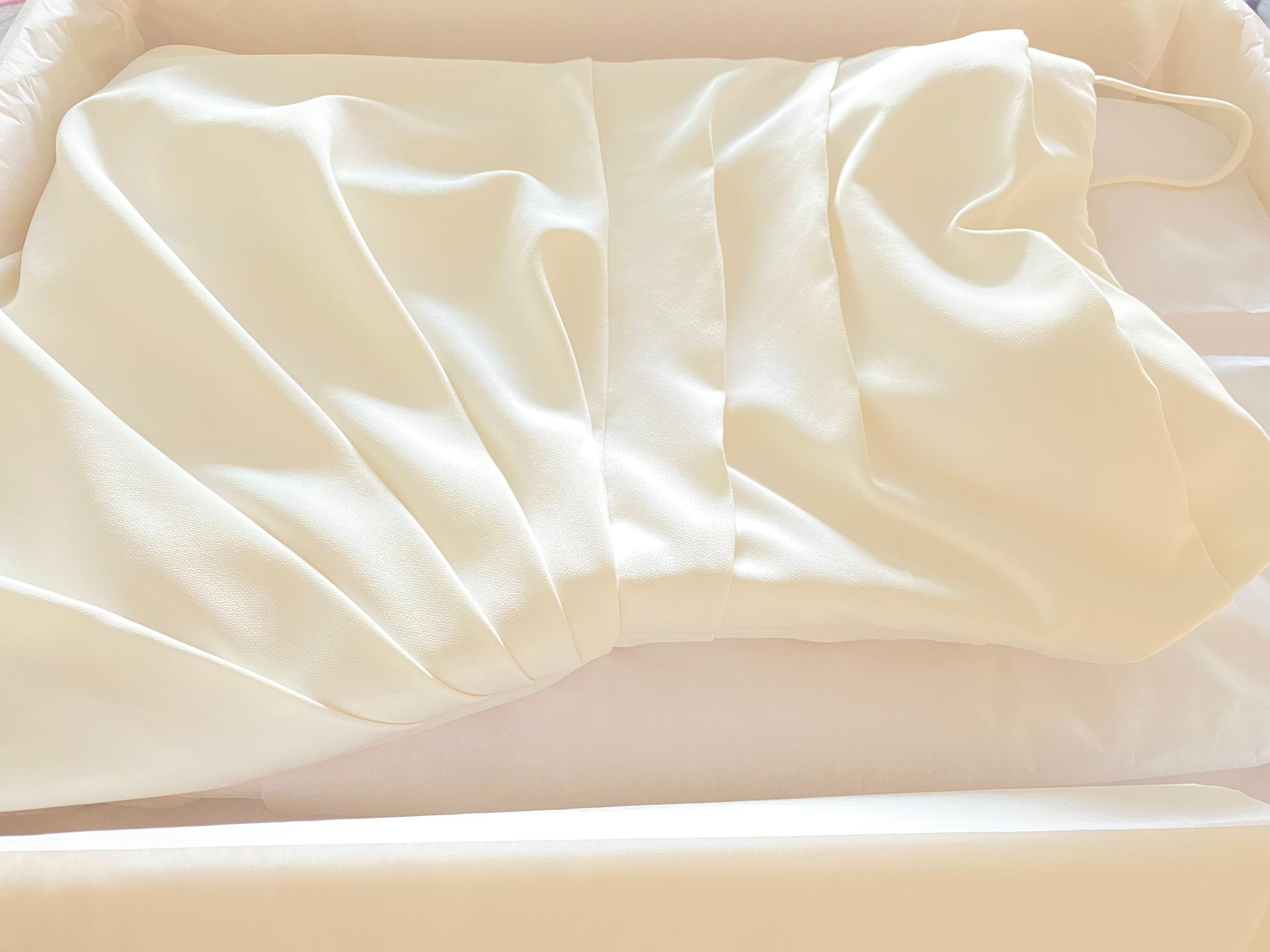 Cream-colored dress folded in a white storage box; the dress has pleated detail on the bodice and spaghetti straps.