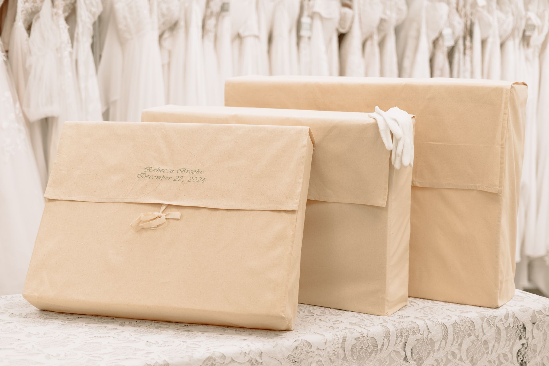 Three wrapped packages on a table. Beige paper covers, one tied with twine. Wedding dresses in background.