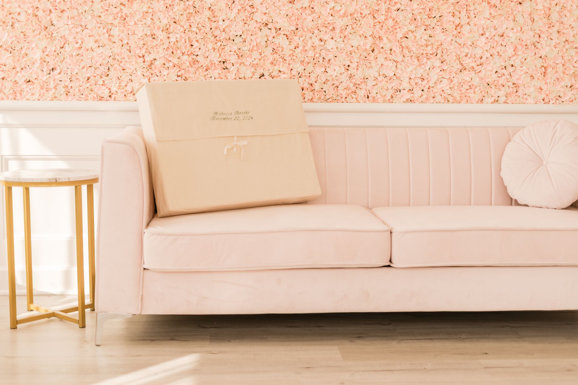 Pink velvet couch with large box, next to a gold side table against a textured pink wall.