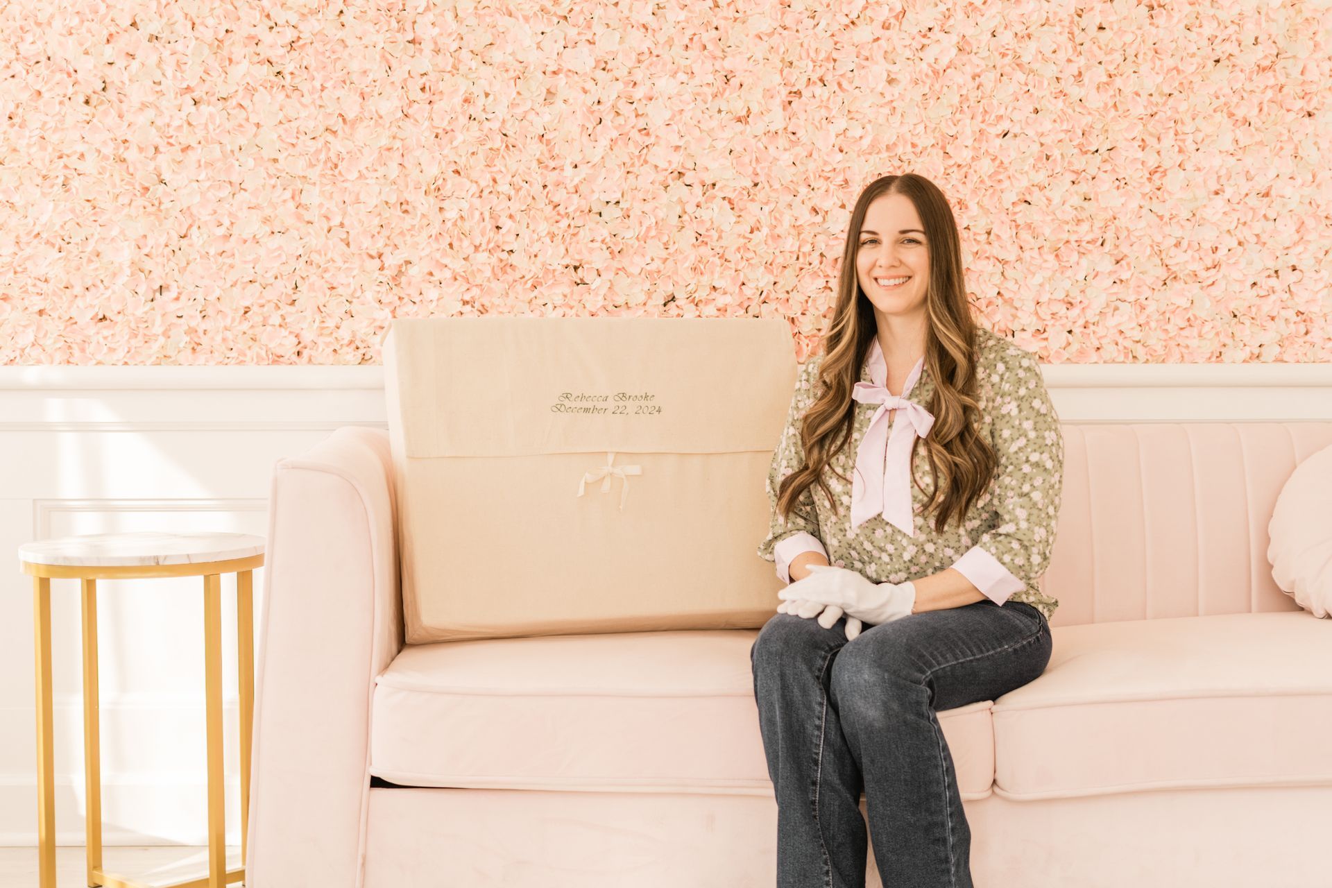 Woman smiling, sitting on a pink couch with a large beige box. Pink floral wall in background.