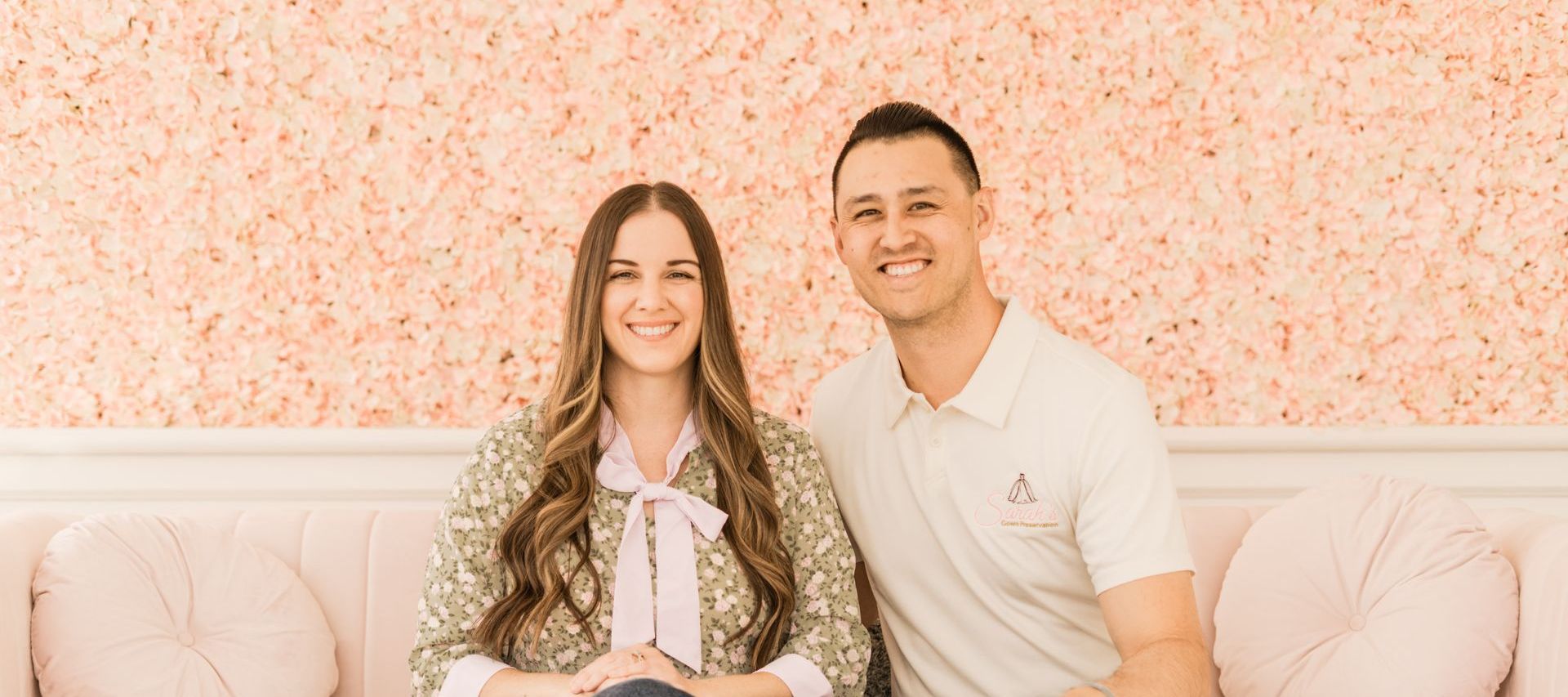 Smiling couple sitting on a pink couch, in front of a pink floral wall.