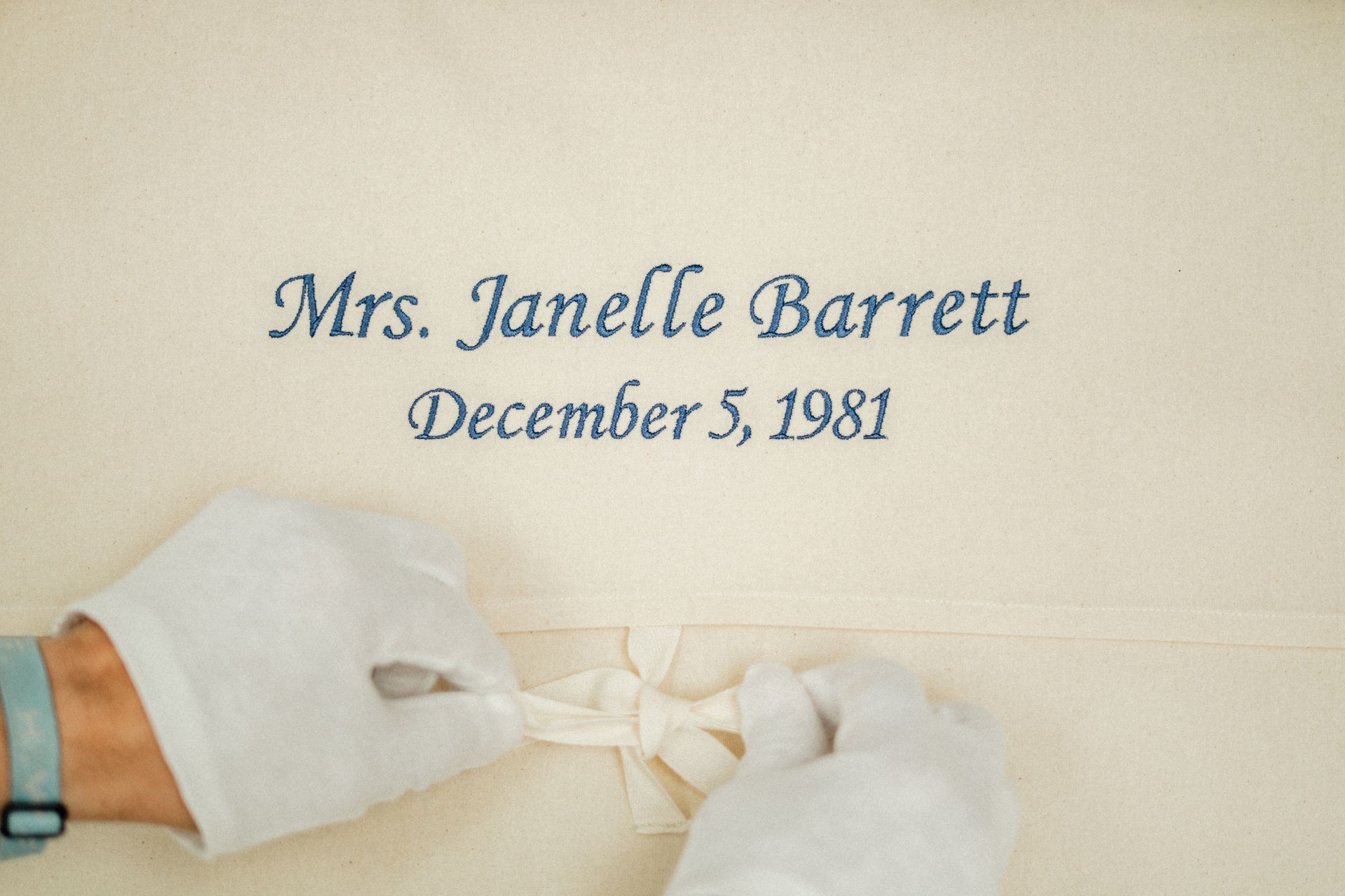 Hands in white gloves untie a ribbon below the embroidered text: 