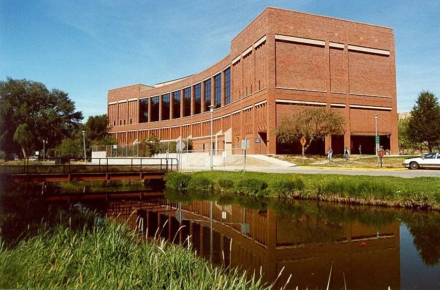 Curved brick building with reflective glass windows stands by a serene pond under a clear blue sky.