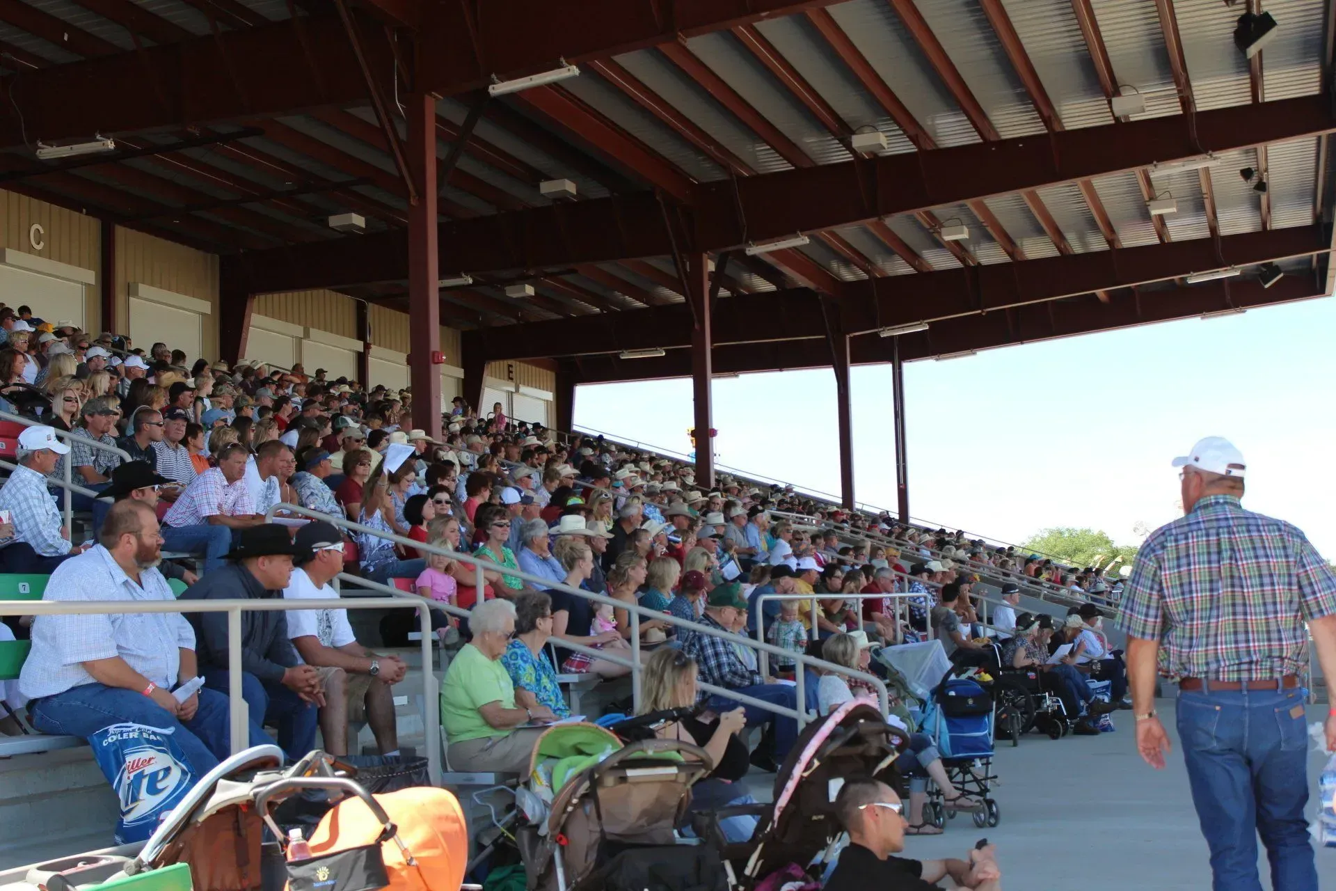 A large crowd sits under a covered bleacher area at an outdoor event. 