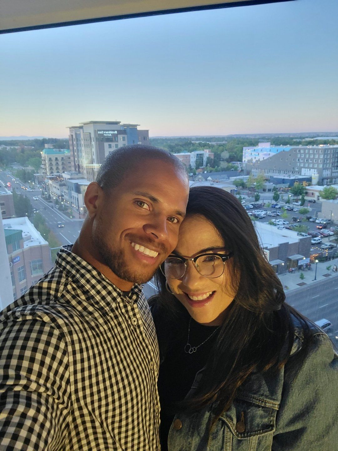 A man and a woman are posing for a picture in front of a window overlooking a city.