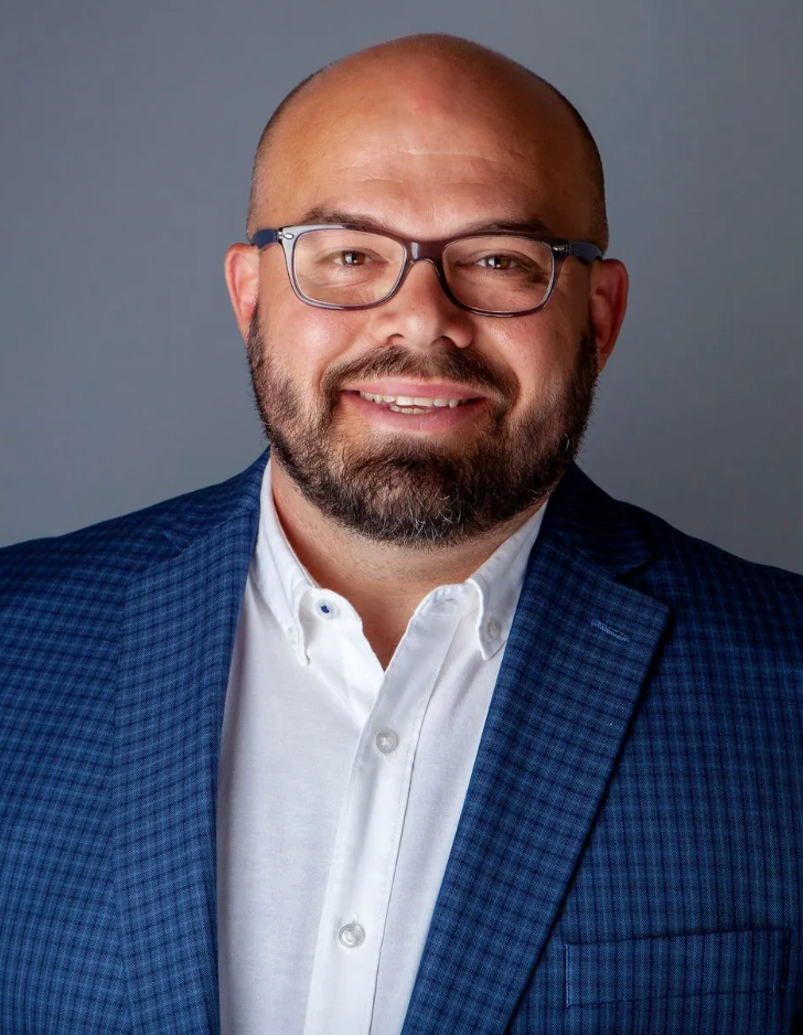 A bald man with a beard and glasses is wearing a blue suit and white shirt.