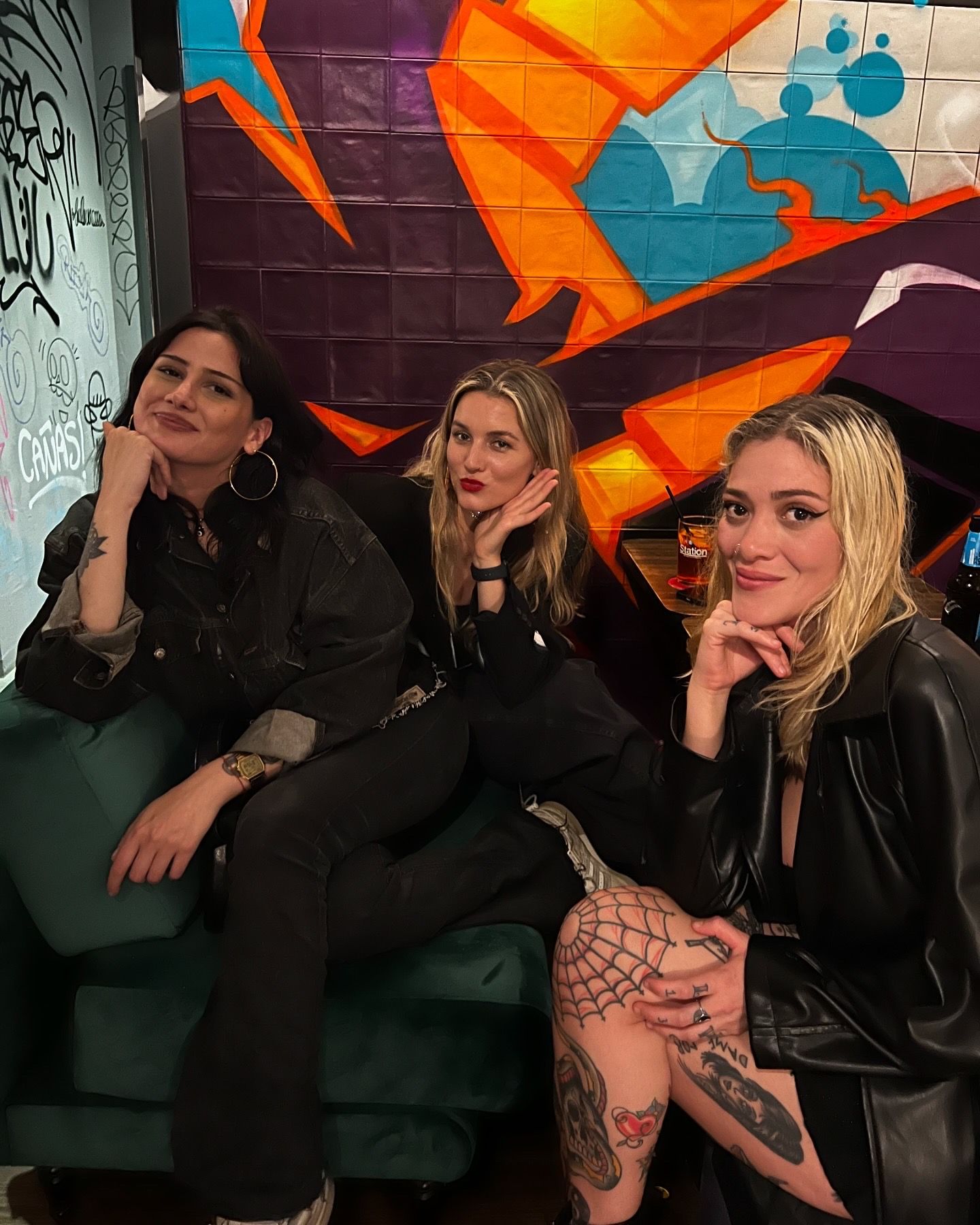 Three women are sitting on a green couch in front of a graffiti wall.