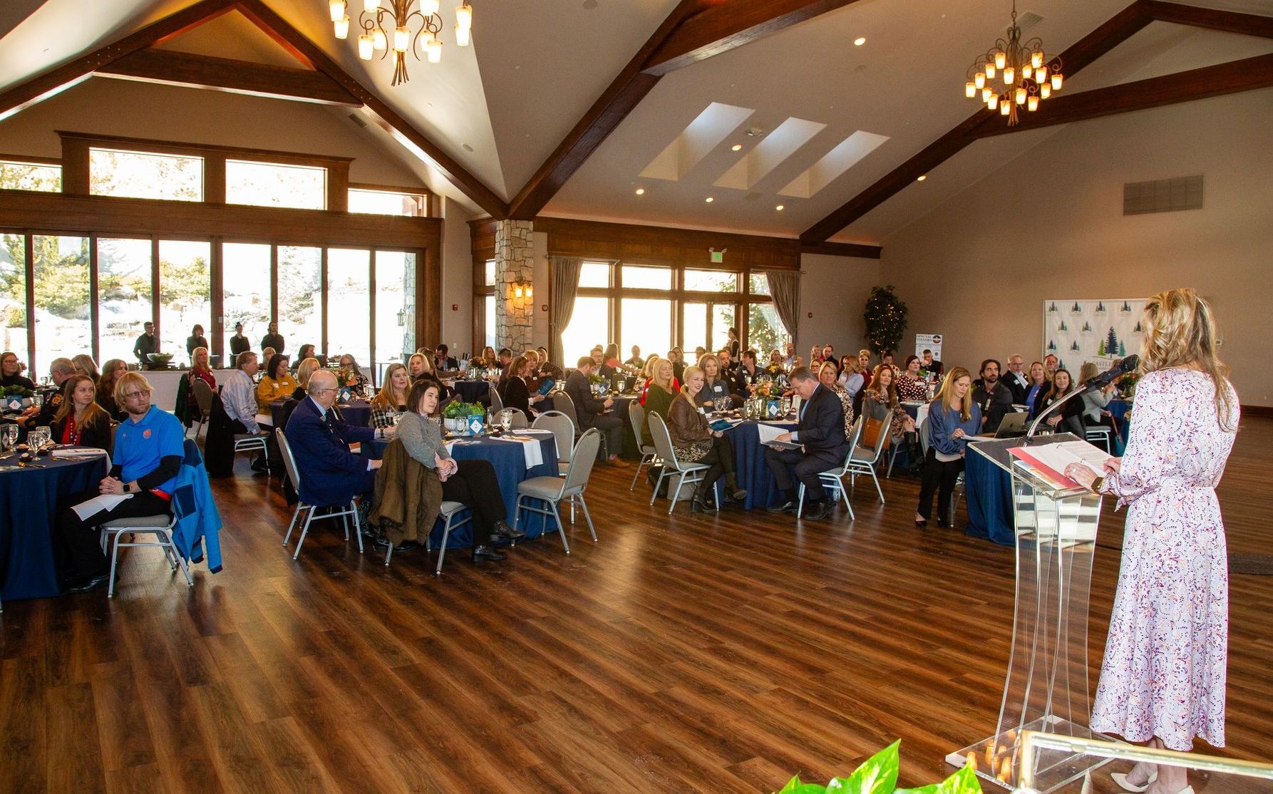 A woman is standing at a podium in front of a large room filled with people sitting at tables.