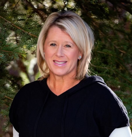 A woman in a black sweater is smiling in front of a tree.