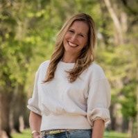 A woman in a white sweater and jeans is smiling in a park.