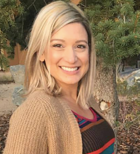 A woman in a tan sweater is smiling in front of a tree.