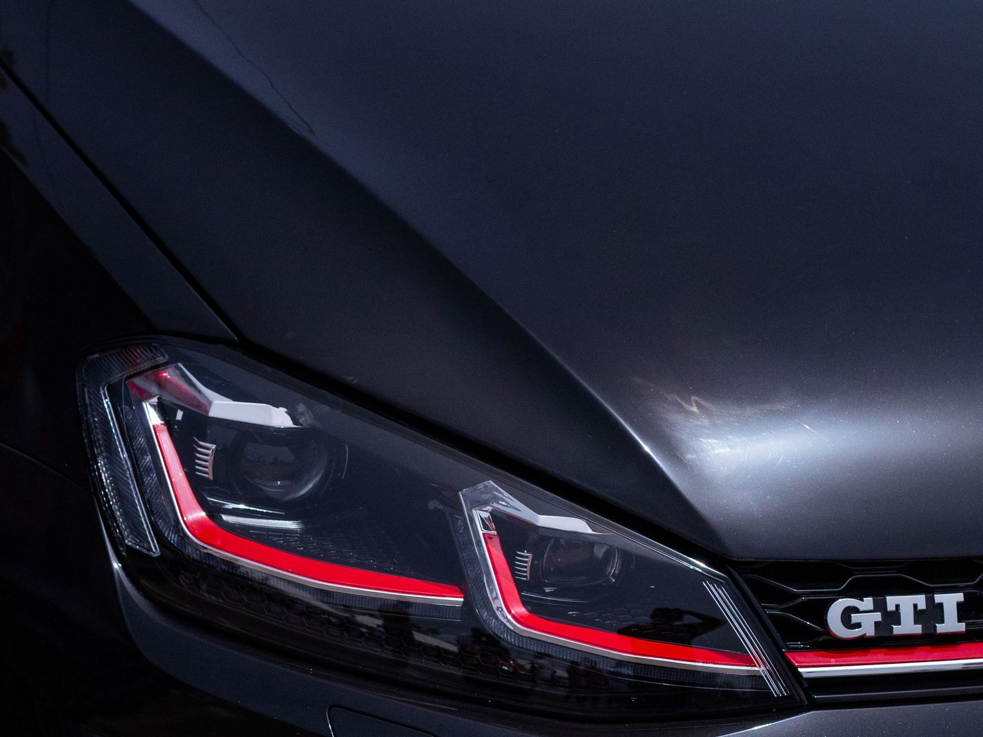 Close-up of a dark grey Volkswagen Golf GTI headlight with distinct red accent lighting and the GTI emblem on the grille.