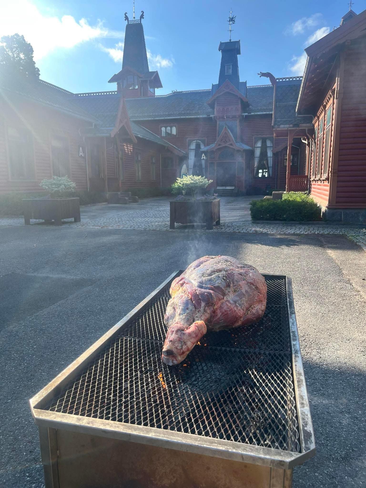 En stor stek som grilles foran en rød trebygning med utsmykket arkitektur under en solrik himmel.