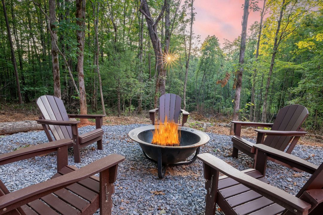 Circular fire pit surrounded by wooden chairs on a gravel patio in a wooded sunset clearing